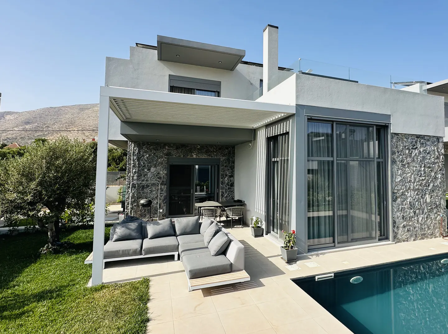 Modern home exterior with a pool, gray stone accents, and a covered patio with a gray sectional sofa. A mountain is visible in the background.