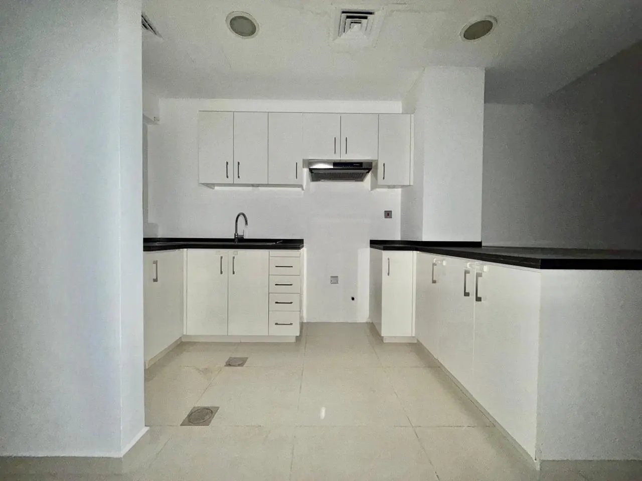 A bright, empty kitchen with white cabinets, black countertops, and light tile flooring.
