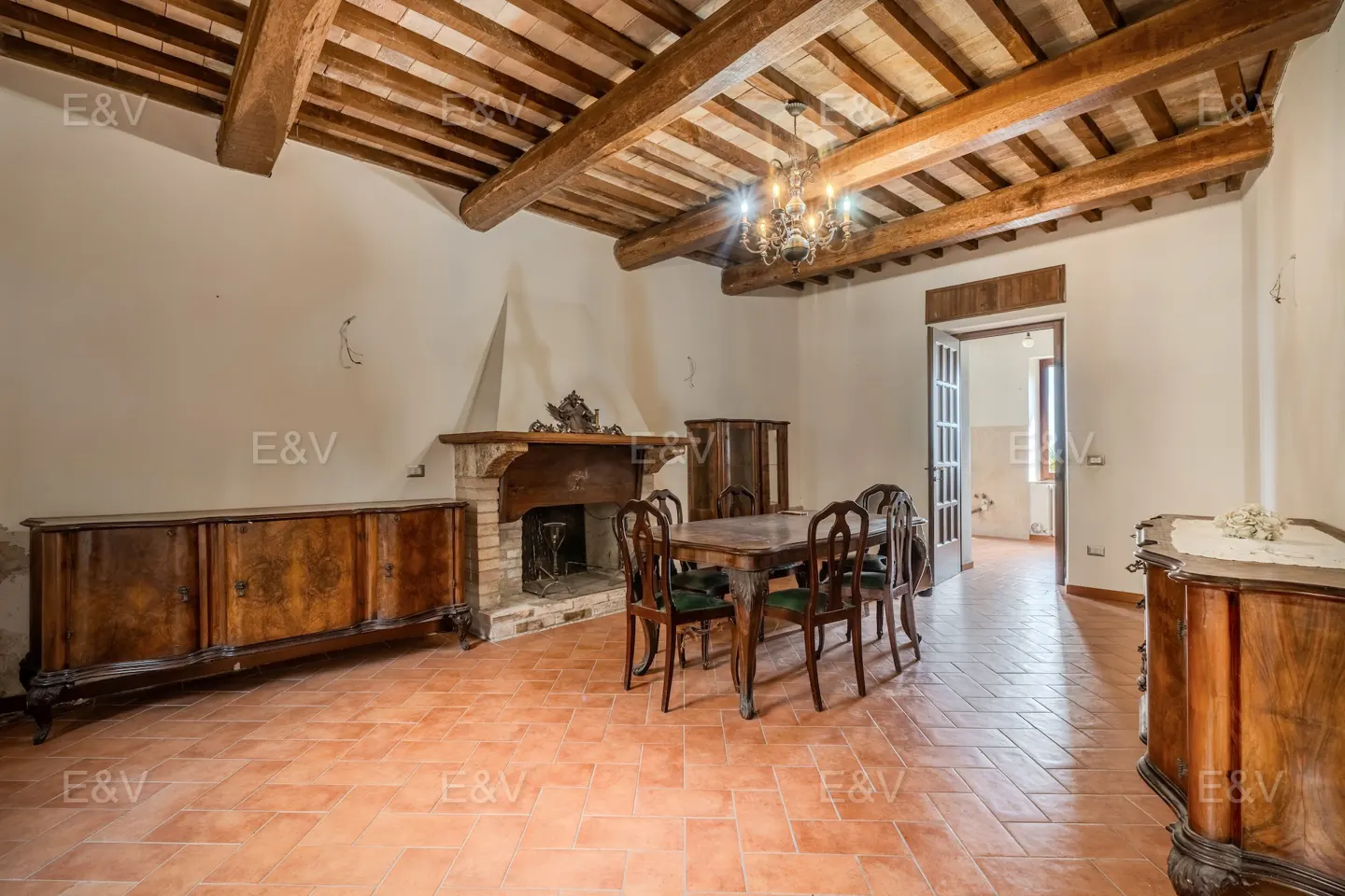 Interior view of a dining room with a fireplace, wooden table and chairs, and terracotta tile flooring.