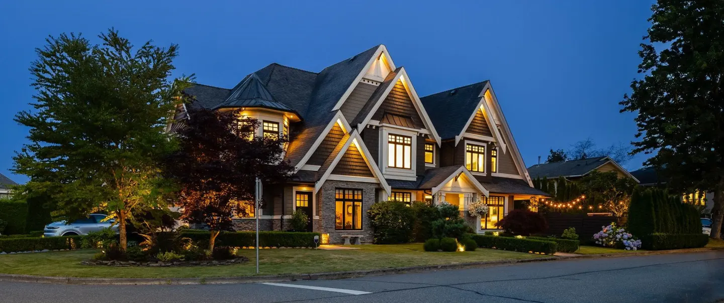 A two-story house with brown siding and a gray roof at dusk. Lights are on inside.