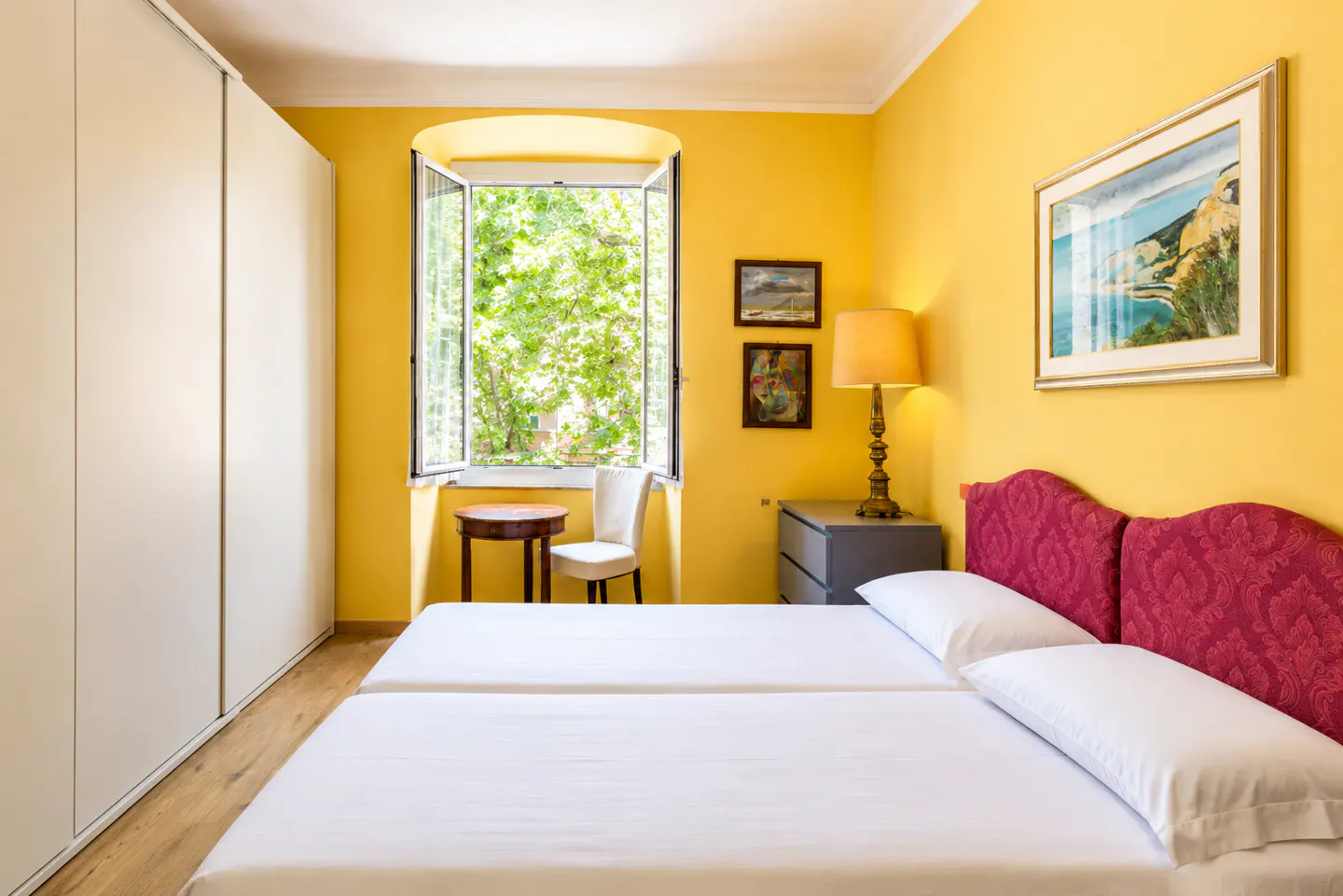 Bright yellow bedroom with two twin beds, white linens, and red patterned headboards. Open window with green trees outside. Closet on the left.