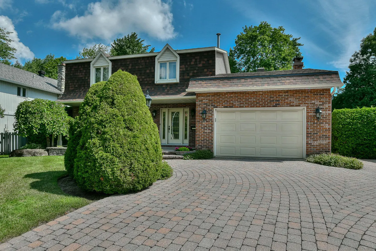 A two-story brick house with a brown roof, white garage door, and a circular brick driveway.