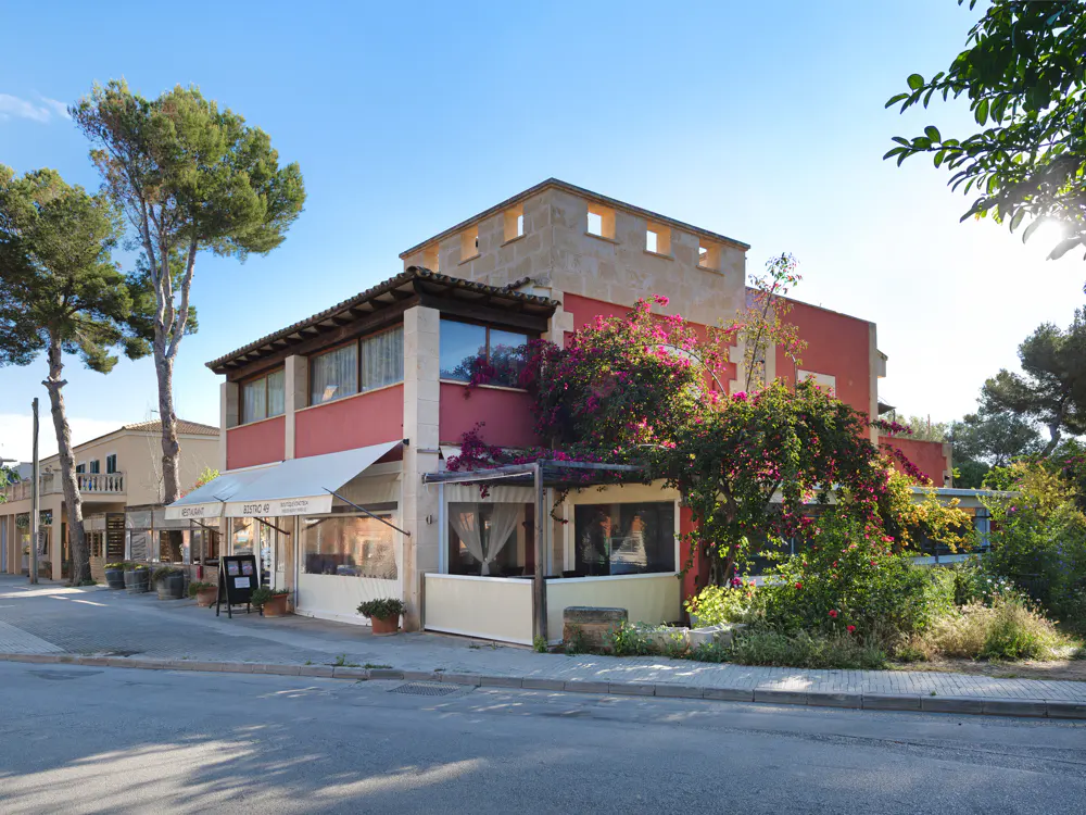Exterior view of a two-story building with a red facade and stone accents, featuring a restaurant with awnings and lush greenery.