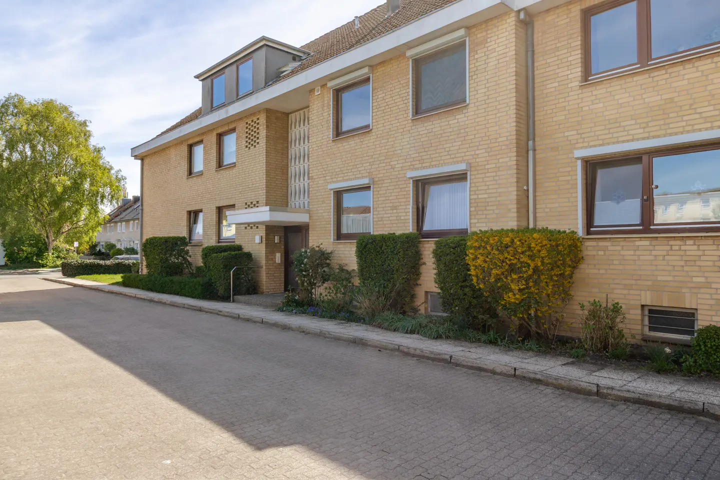 Exterior view of a two-story yellow brick apartment building with brown window frames and green bushes.