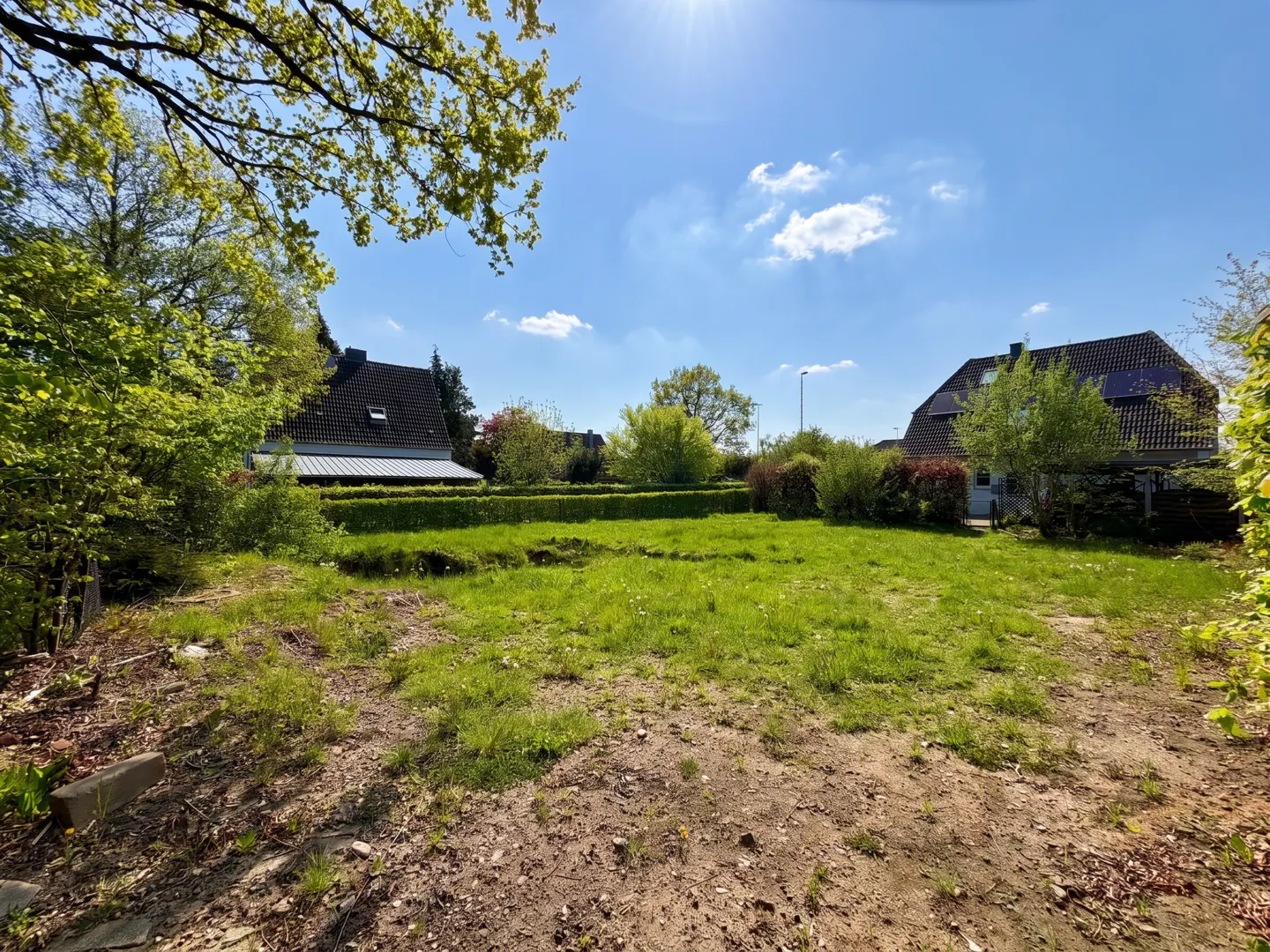 Vacant lot with green grass and dirt patches, surrounded by hedges and trees under a sunny blue sky. Houses are visible in the background.