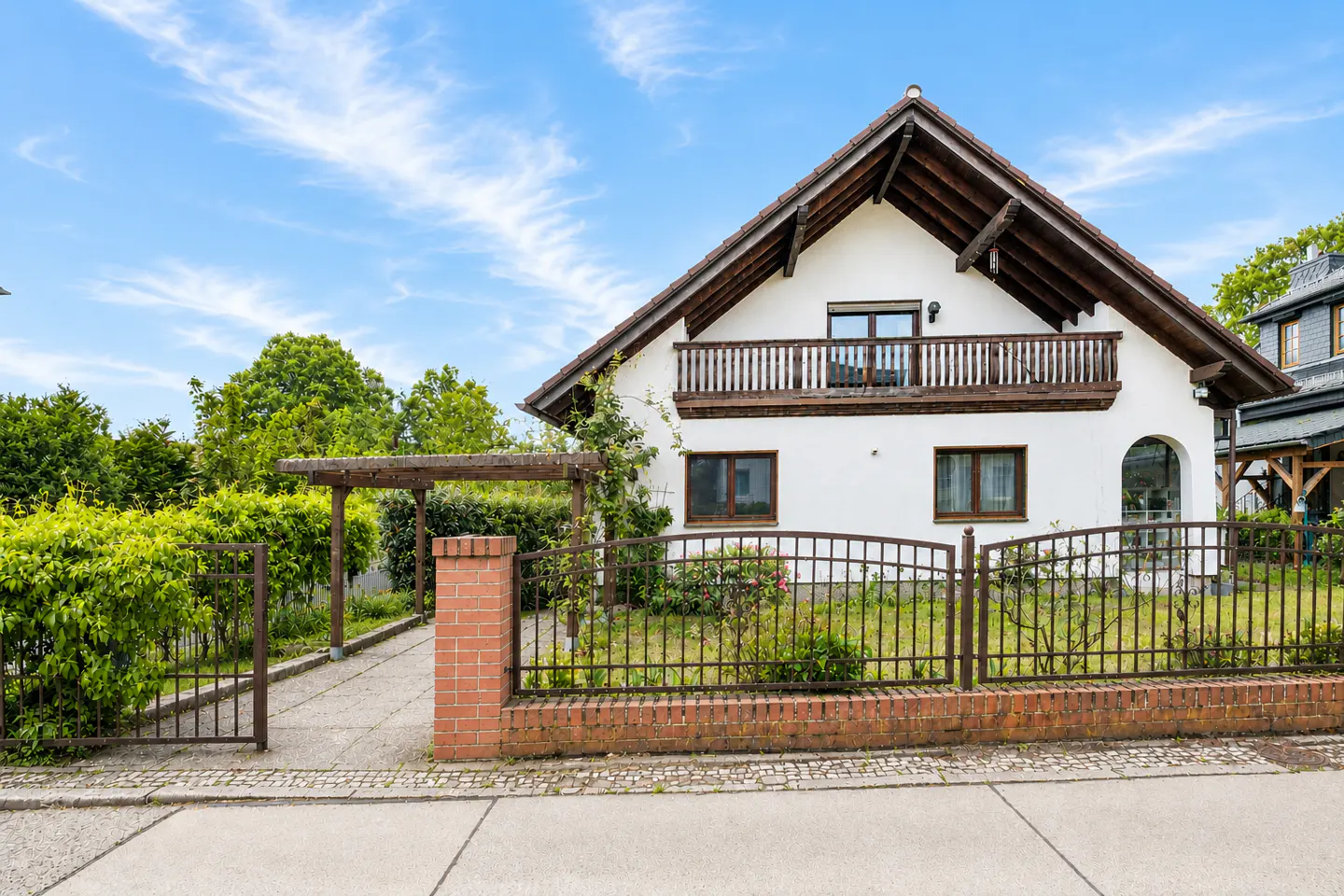 Two-story white house with brown trim, a balcony, and a metal fence, under a blue sky with wispy clouds.