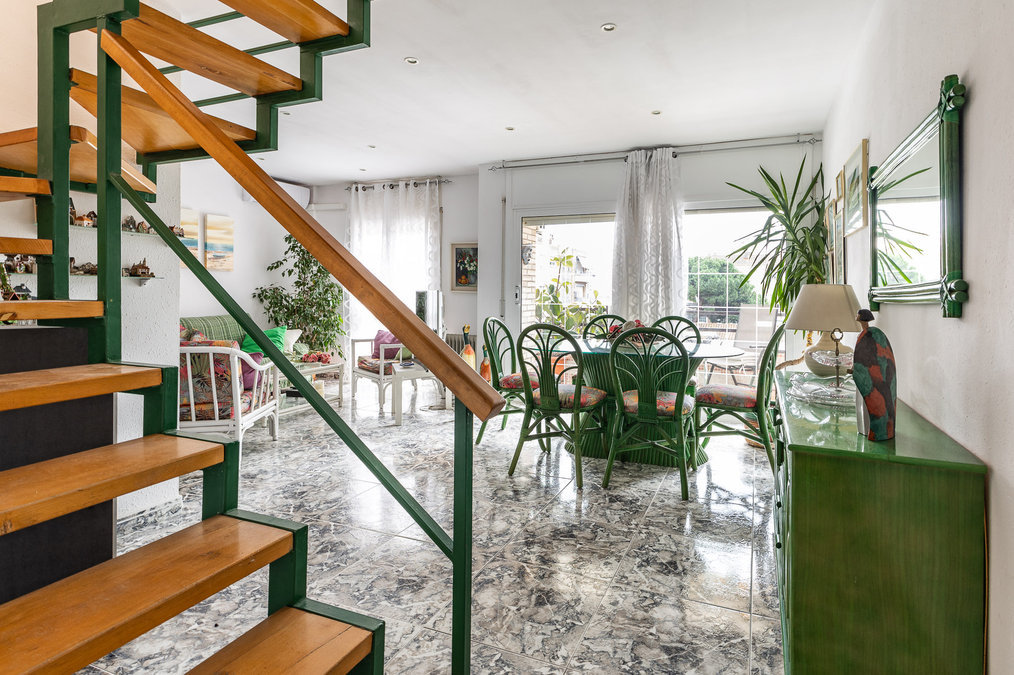 Open-plan living room with marble floors, a green dining set, and a wooden staircase with green railings. [AI-generated text]