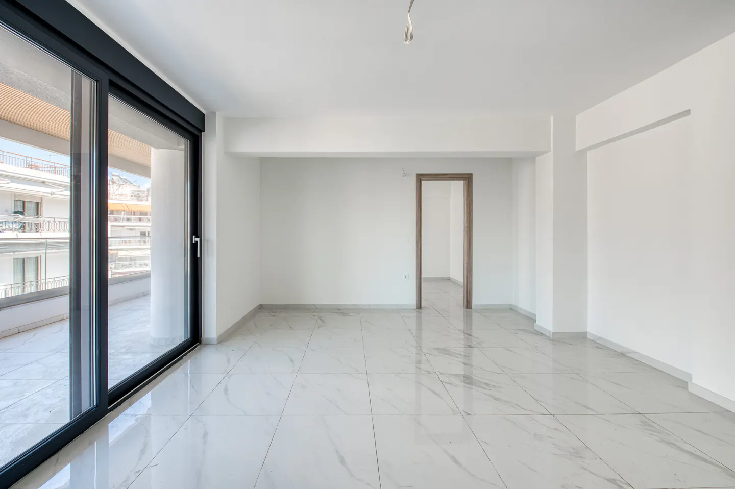 Bright, empty room with white walls, marble tile floor, and black-framed sliding glass doors to a balcony. A doorway leads to another room.