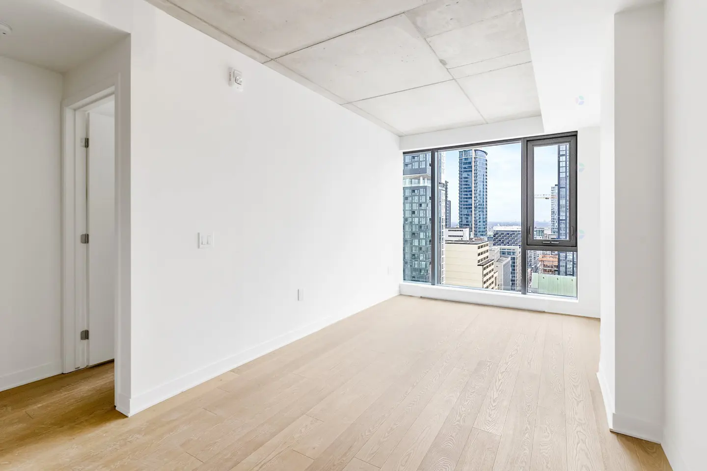 Empty modern room with white walls, light wood floors, exposed concrete ceiling, and large windows with city views.