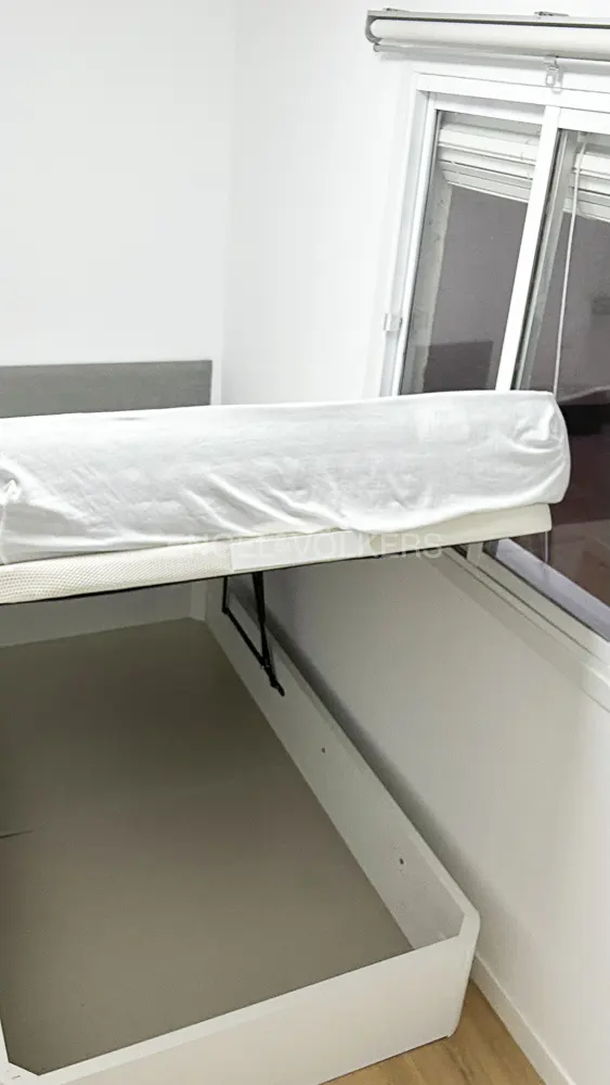 A white storage bed with the mattress lifted, revealing an empty interior compartment next to a window in a white room.