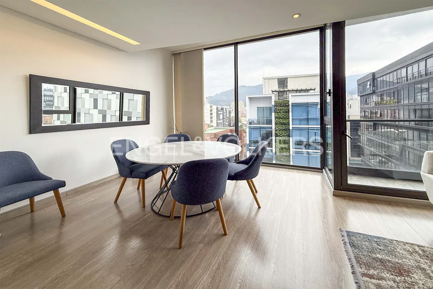 Bright dining room with wood floors, a round marble table, and blue chairs. Large windows offer city views.