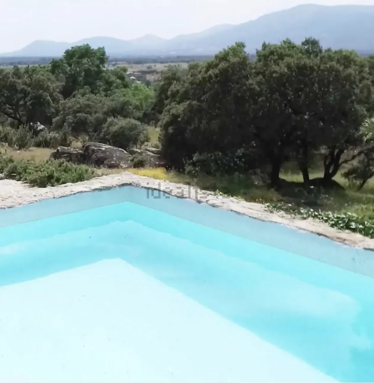 A light blue swimming pool with a stone border overlooks a green, tree-filled landscape and distant mountains.