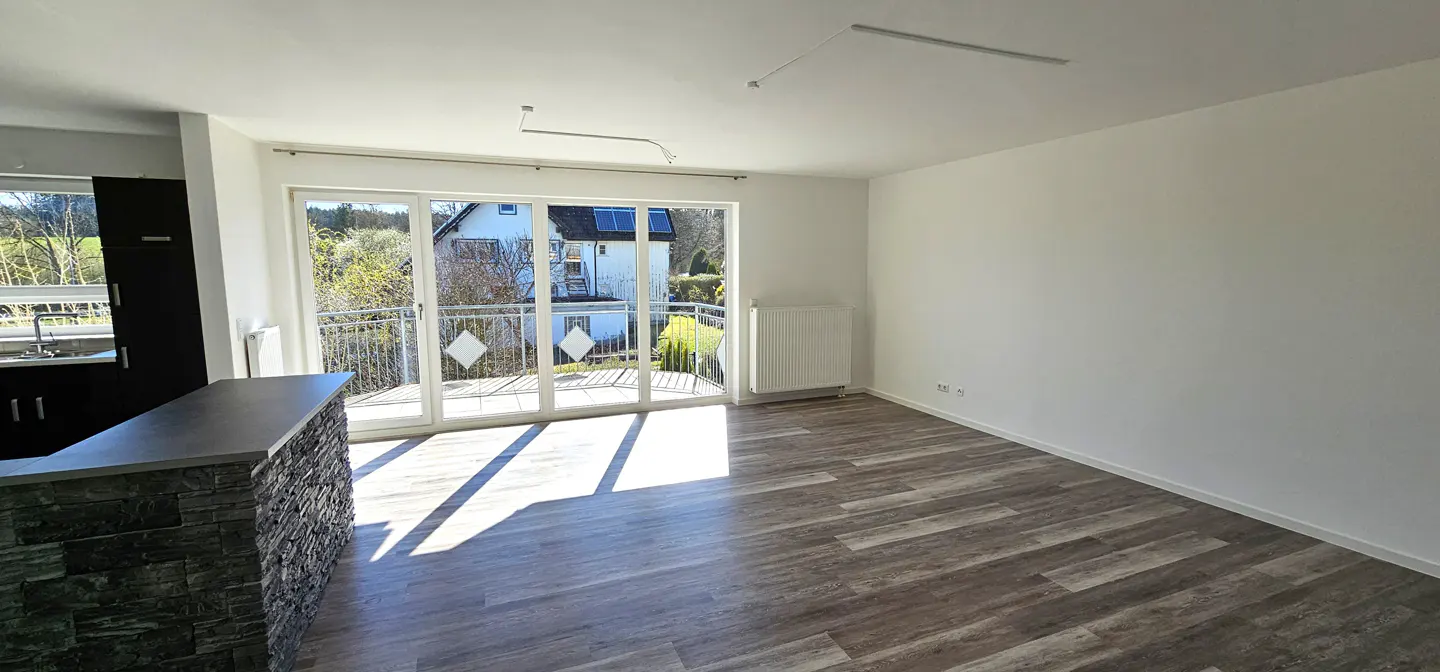 Bright, empty room with wood-look floors, white walls, and a stone-clad kitchen island. Balcony doors open to a view of a house and trees.