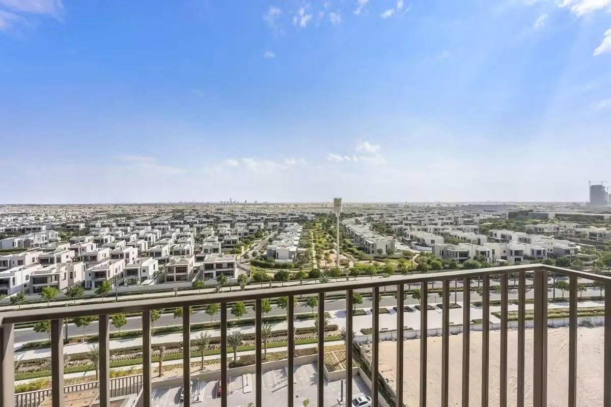 View from a balcony with a metal railing overlooking a residential area with white houses, green trees, and a blue sky.