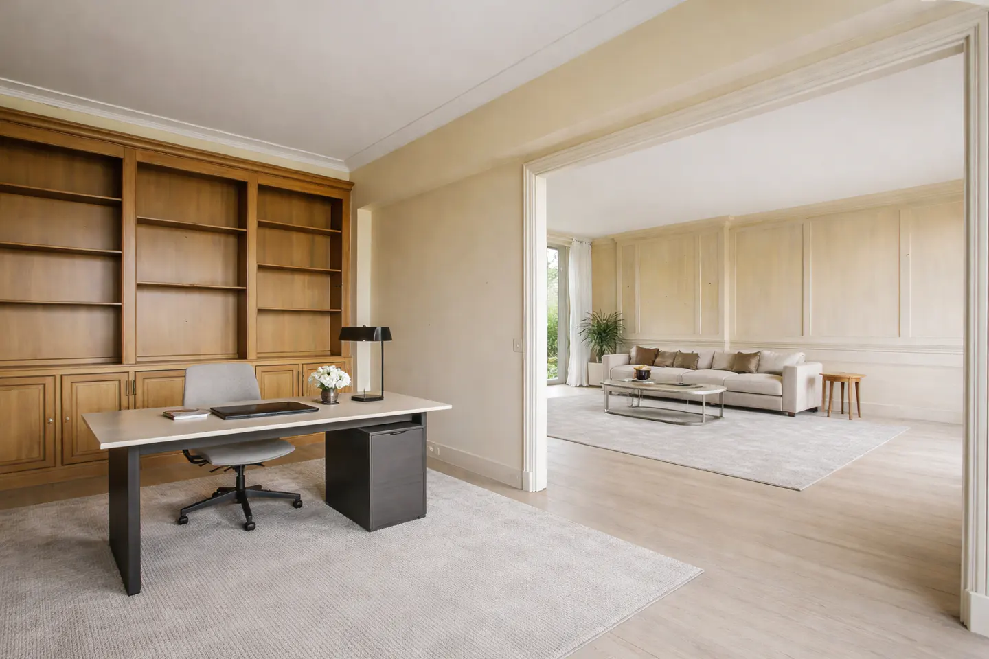 A bright home office with a desk, chair, and built-in wooden bookcase, leading to a living room with a sofa.