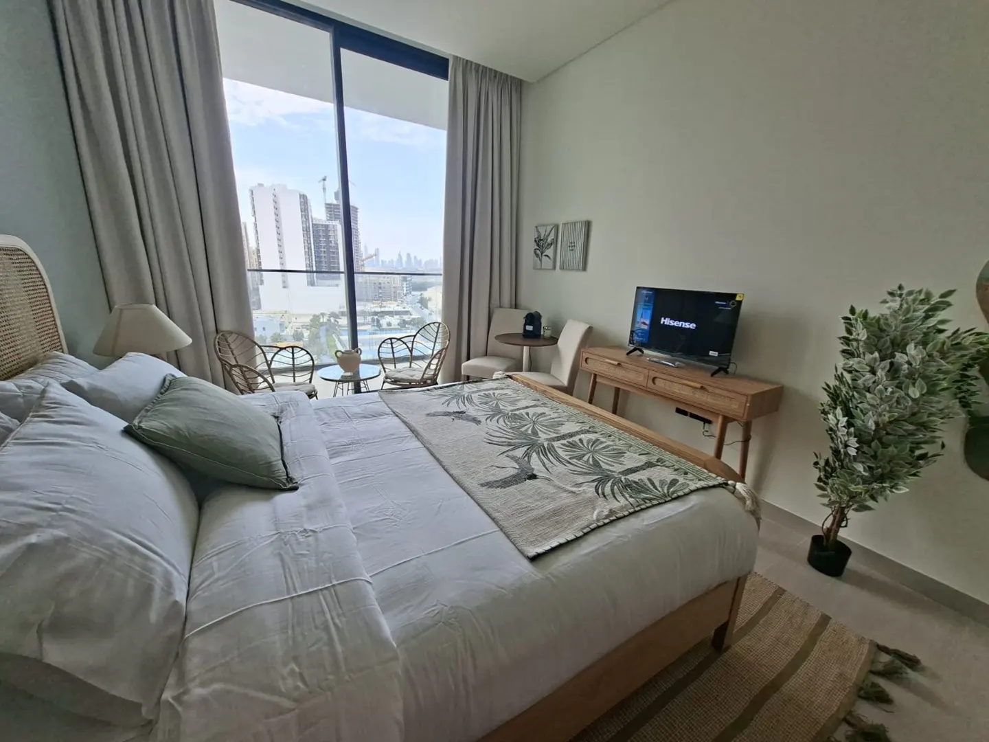 Bedroom with a large bed, white linens, a palm-leaf patterned throw, and a city view from the balcony. A TV sits on a wooden console.