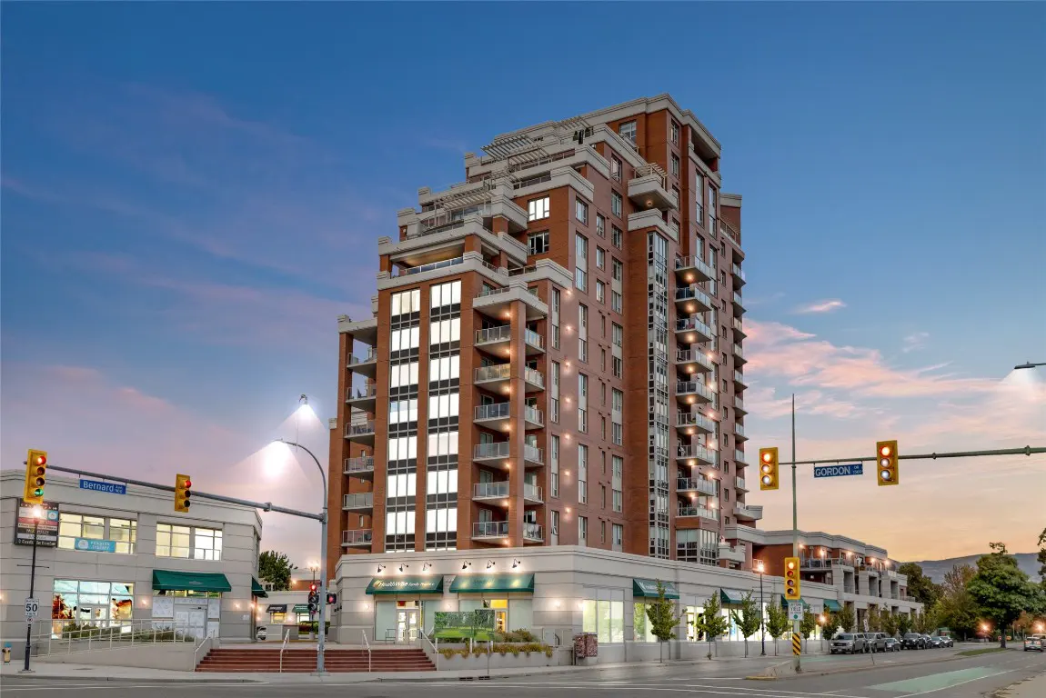A tall, red brick condo building with balconies, street-level shops, and a blue sky at dusk.
