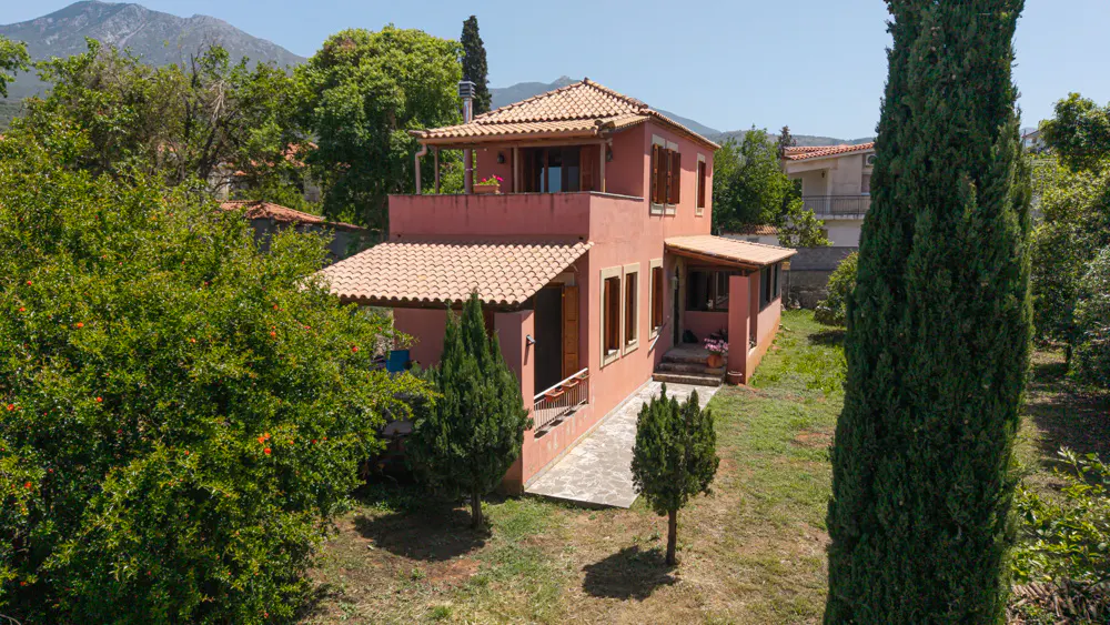 Two-story peach house with a tile roof, brown shutters, and a balcony, surrounded by green trees and a mountain backdrop.