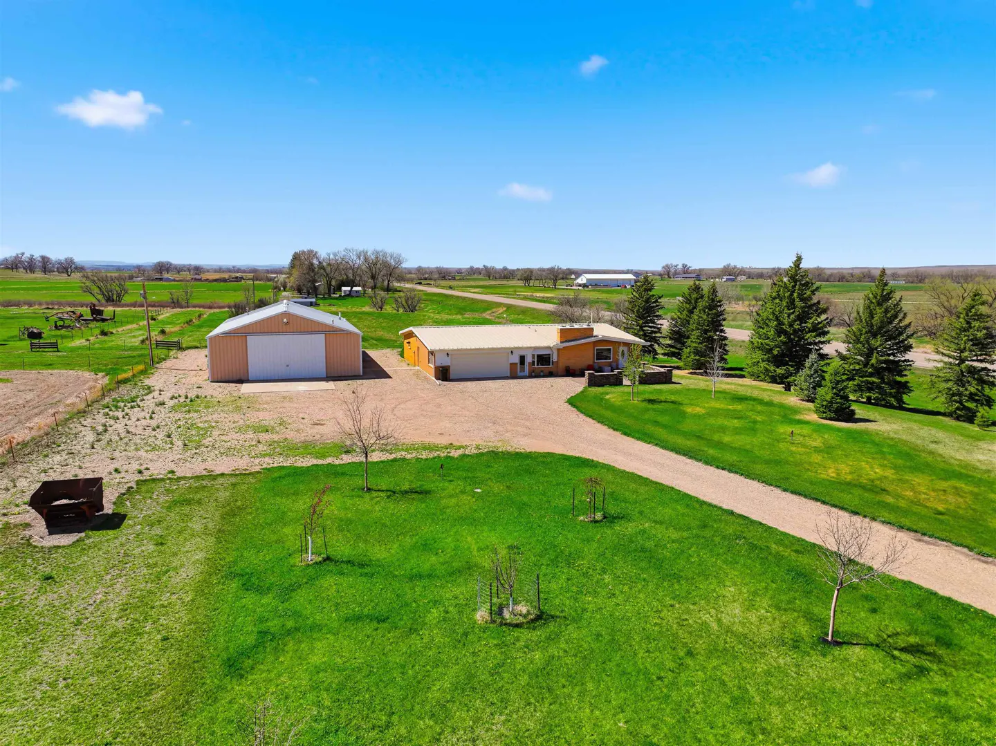 Aerial view of a rural property with a tan house, detached garage, gravel driveway, and green lawn under a blue sky.
