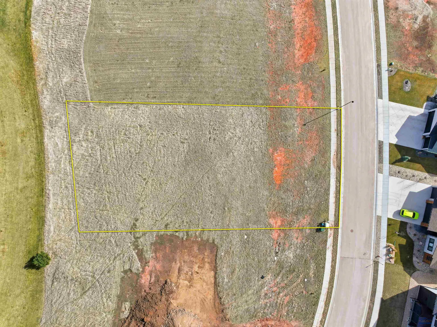 Aerial view of a vacant lot outlined in yellow, adjacent to a street and houses in a residential area.