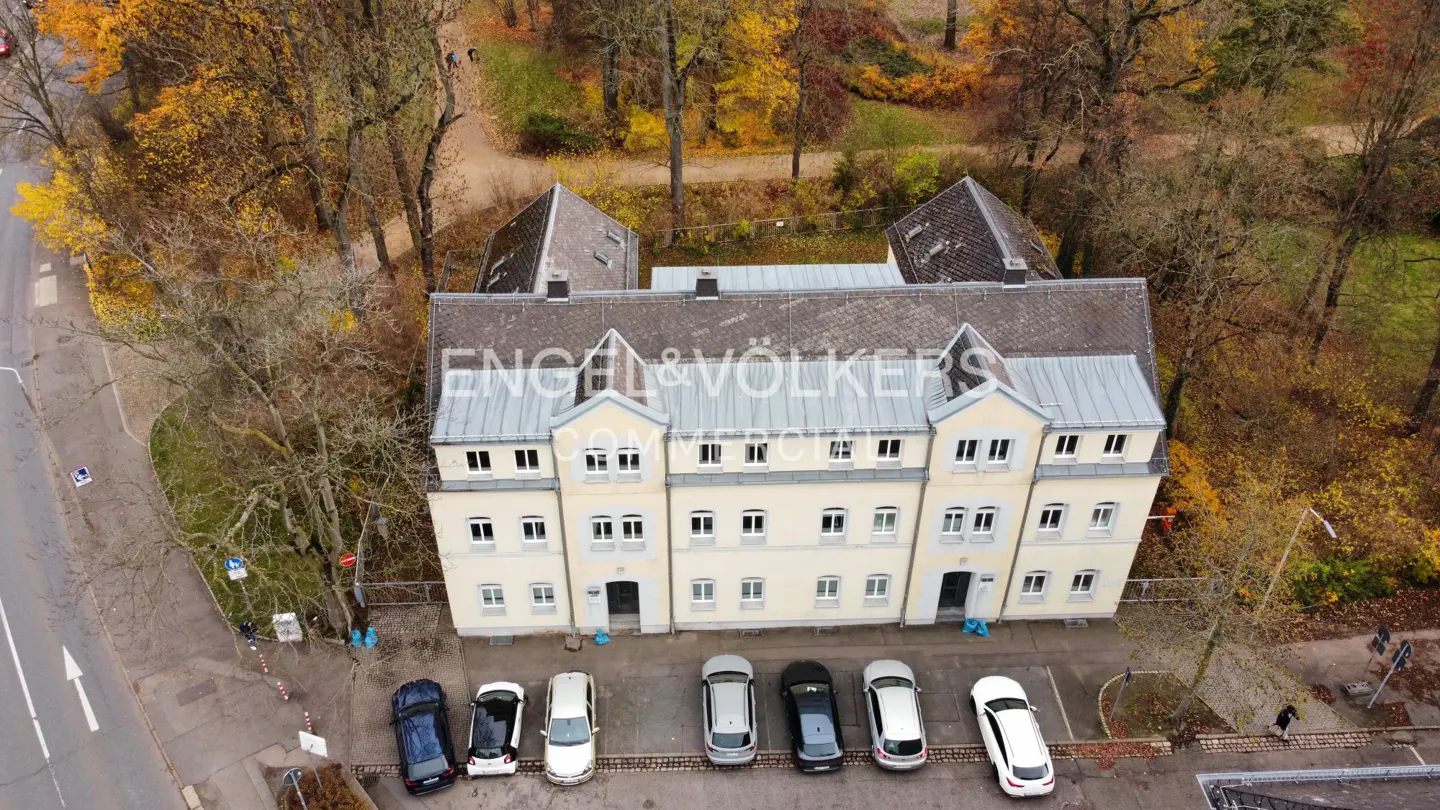 Aerial view of a cream-colored, three-story building with a gray roof, surrounded by trees with autumn foliage and parked cars.