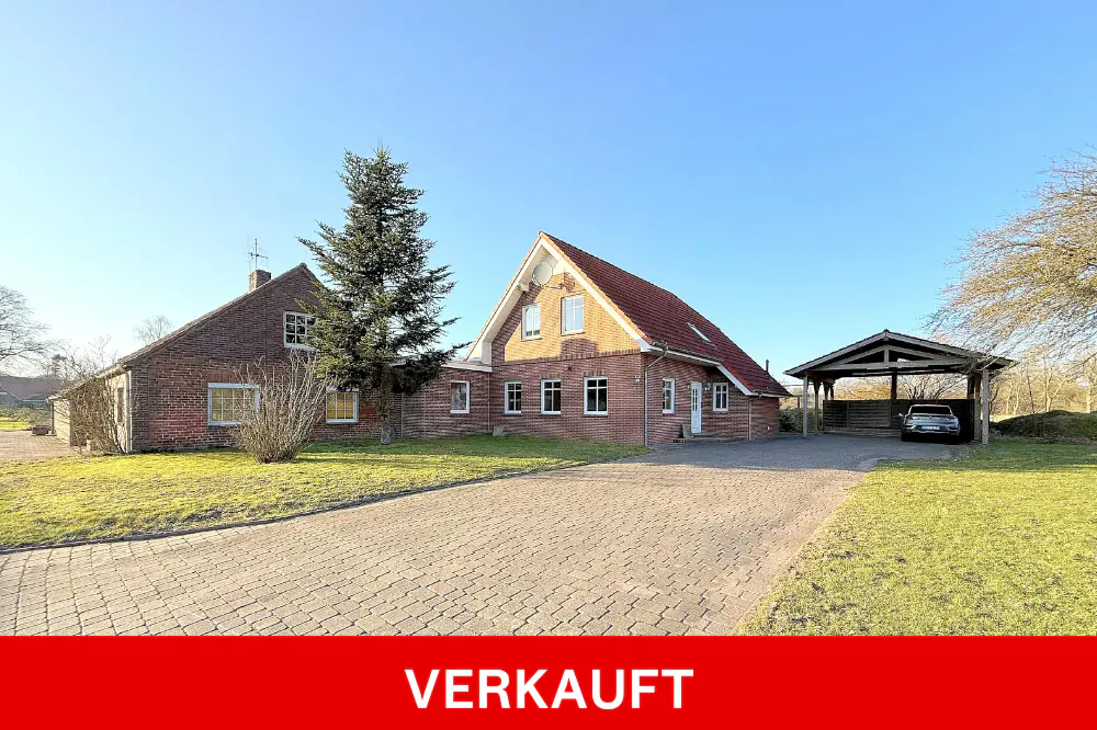Two-story brick house with a red tile roof, a carport, and a paved driveway on a sunny day. "VERKAUFT" is written in white on a red banner.