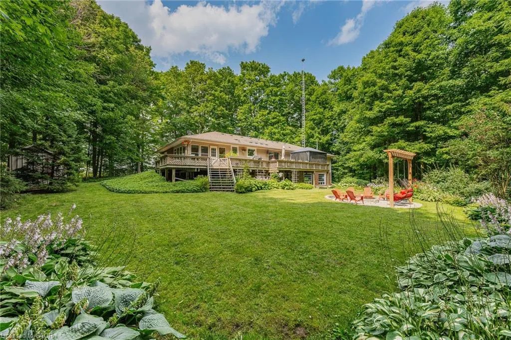 A tan house with a wooden deck overlooks a green lawn, surrounded by trees and a blue sky. A fire pit with orange chairs sits on the lawn.