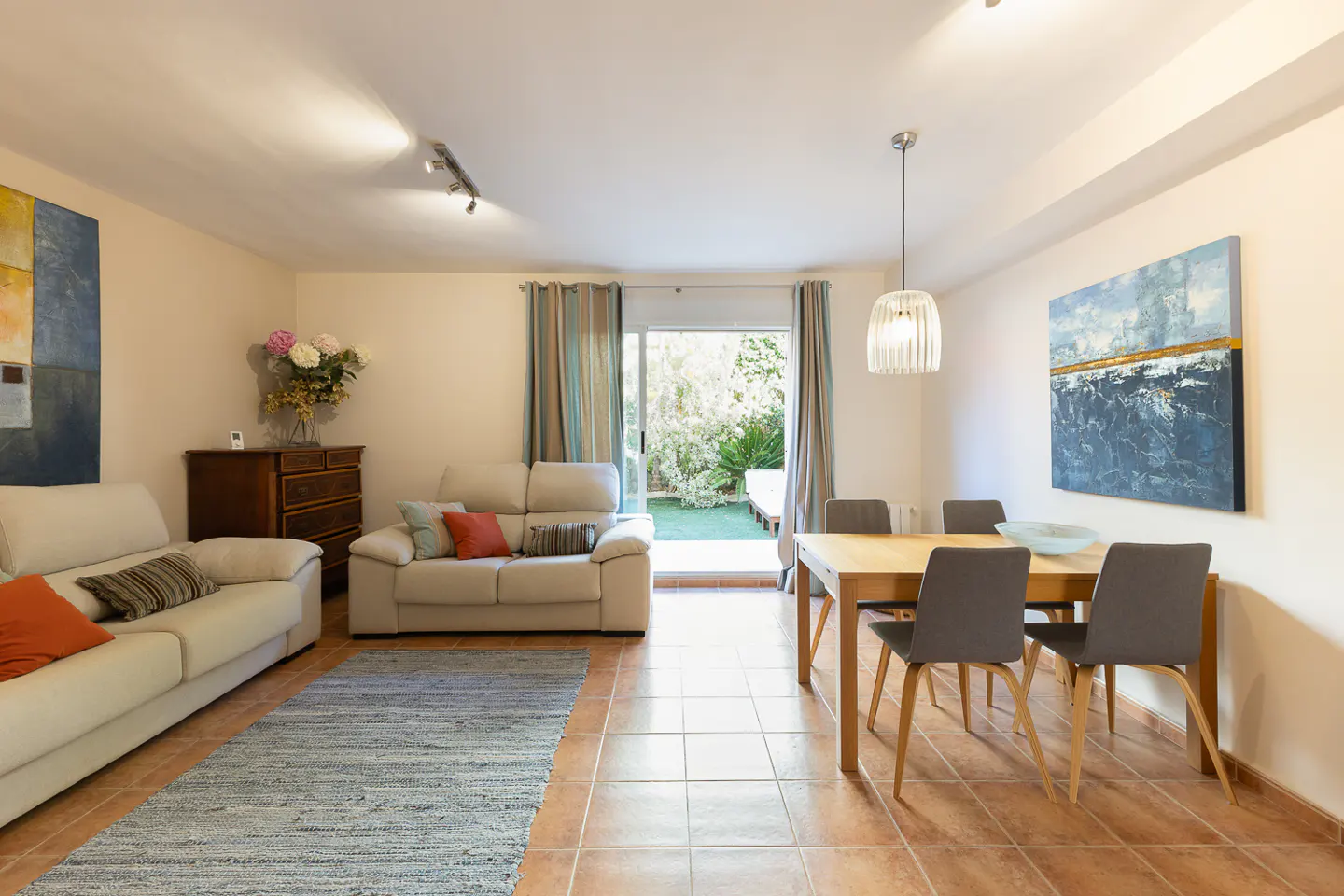 A bright living room with two white sofas, a dining table with four chairs, and a sliding glass door to a garden.