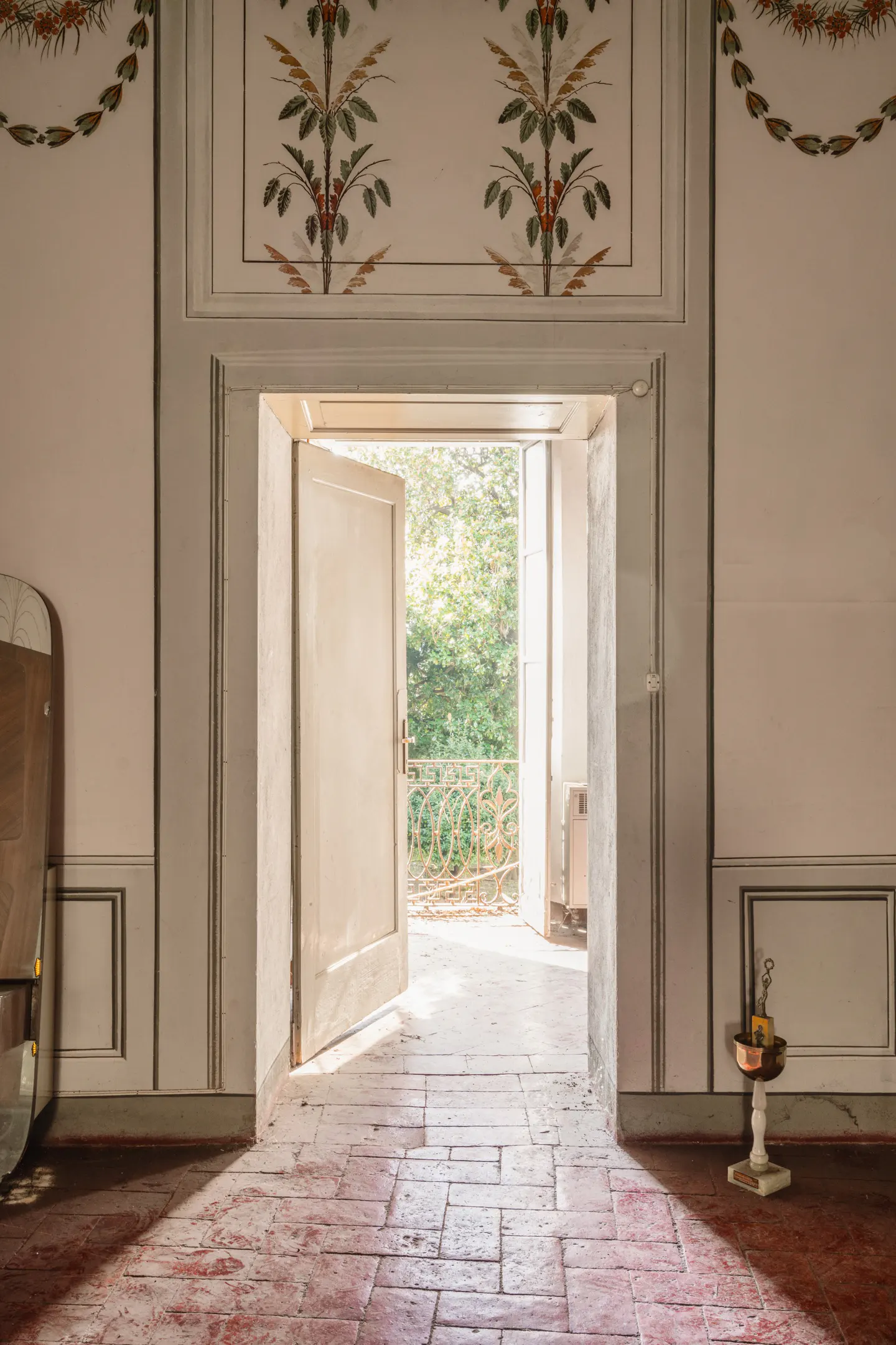 Interior view of a room with red brick floors and white walls. An open door leads to a balcony with trees in the background.