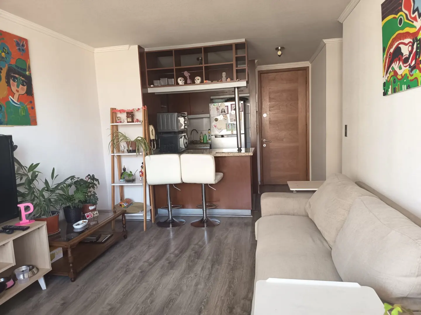 A bright living room with a beige sofa, wood floors, and a kitchen with two white bar stools. Colorful art hangs on the white walls.