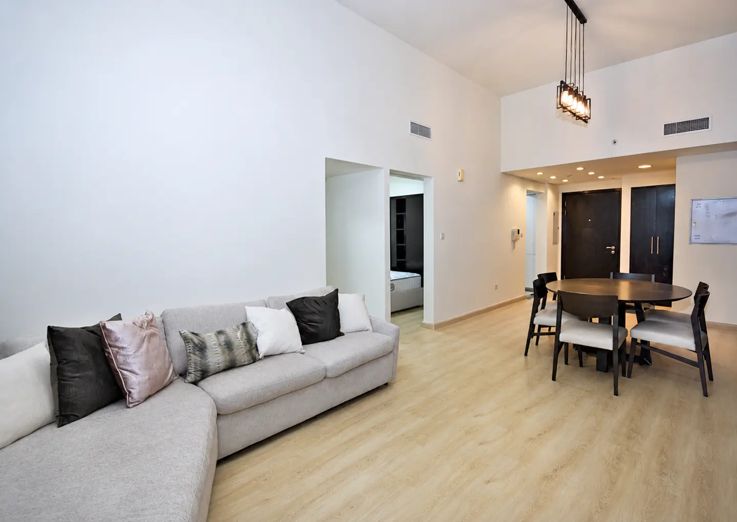 Bright living room with a gray sectional sofa, pillows, and a round dining table with chairs on a light wood floor.