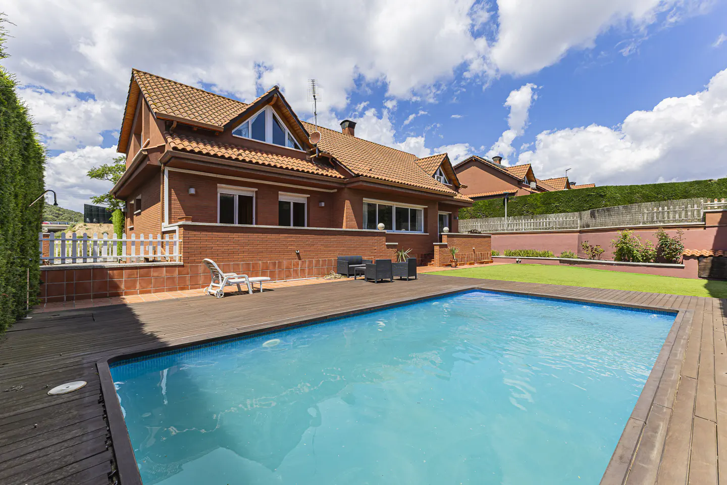 A red brick house with a pool and a wooden deck under a blue sky with white clouds.
