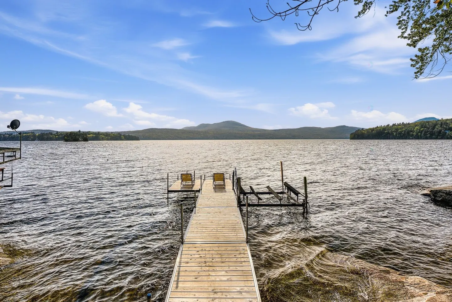 Lake view with a wooden dock leading to two yellow chairs. Mountains and blue sky with clouds in the background.