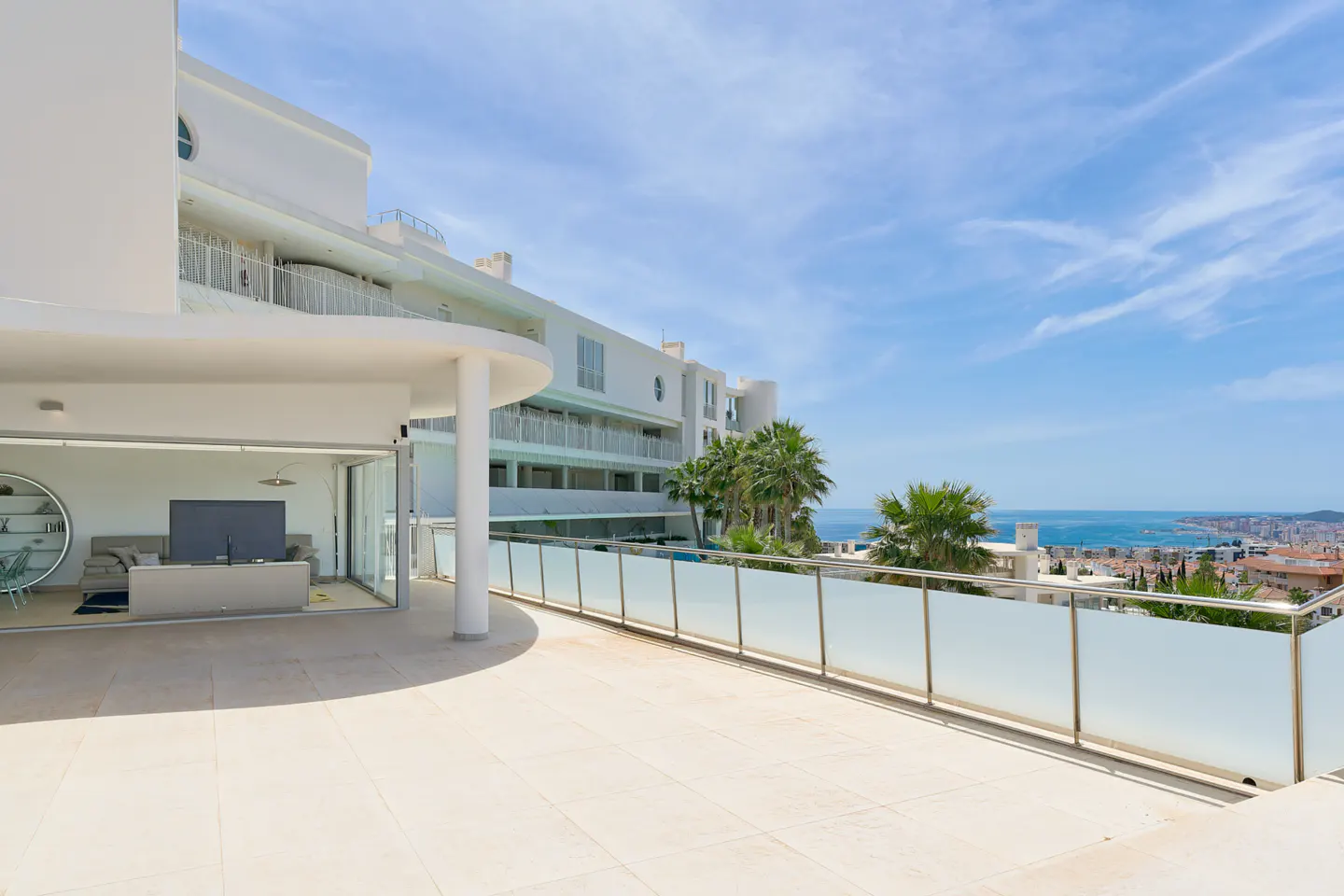 A bright, modern apartment balcony with a frosted glass railing overlooks a coastal city and the sea under a blue sky.