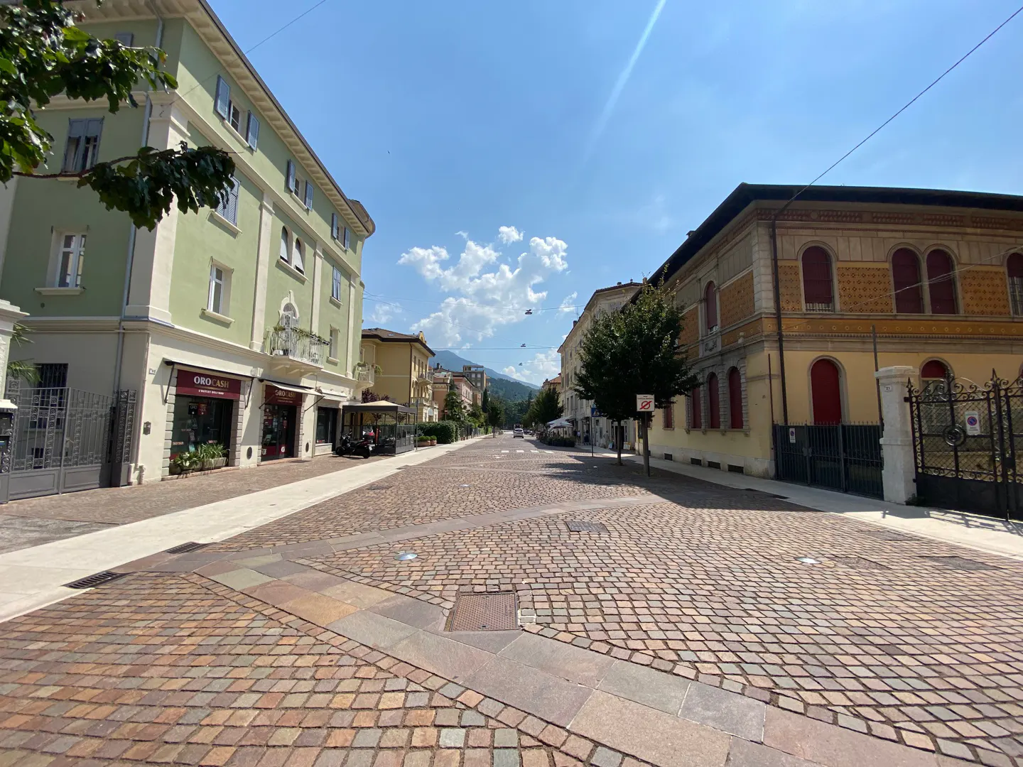 Cobblestone street view in Italy with light green and yellow buildings under a blue sky with white clouds.