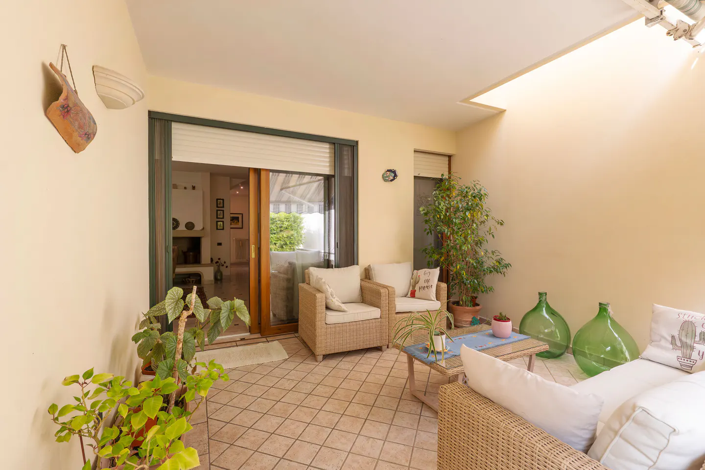 A sunroom with wicker furniture, plants, and a skylight. The walls are cream-colored, and the floor is tiled. A sliding glass door leads to the interior.