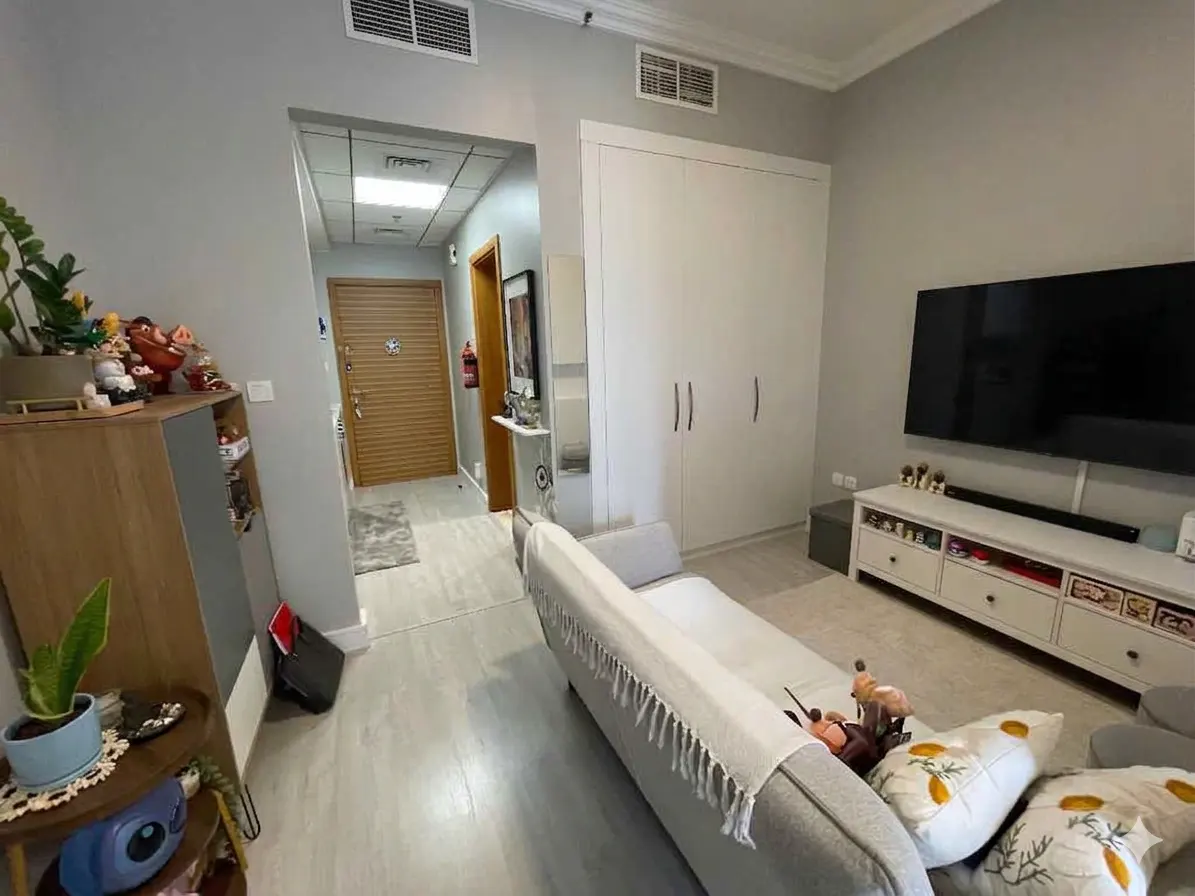 Living room view with gray walls, light wood floors, a gray sofa, and a large TV on a white console. A hallway leads to a wooden door.