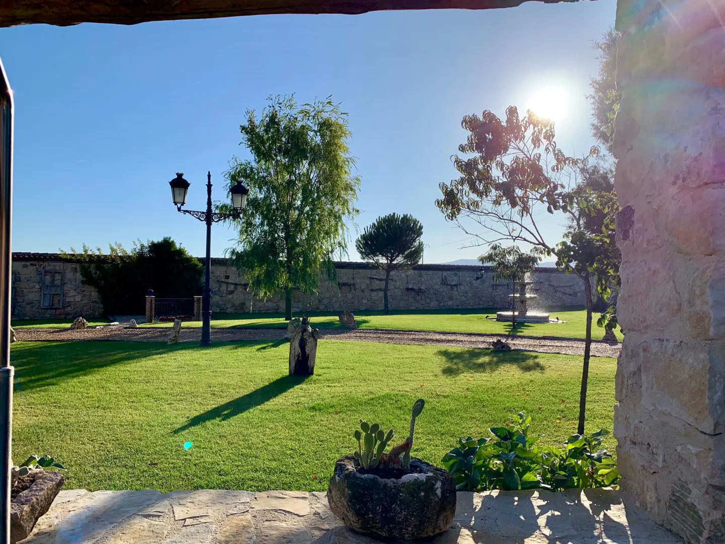 View of a sunny, green garden with a stone fountain, lamp post, and stone wall, framed by a rustic stone porch.
