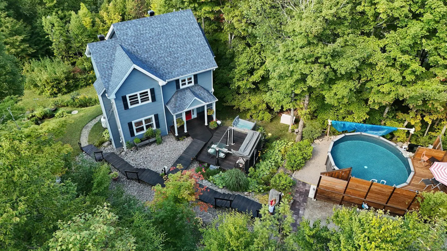 Aerial view of a blue two-story house with a gray roof, black shutters, and a red door, surrounded by lush green trees, a pool, and a hot tub.
