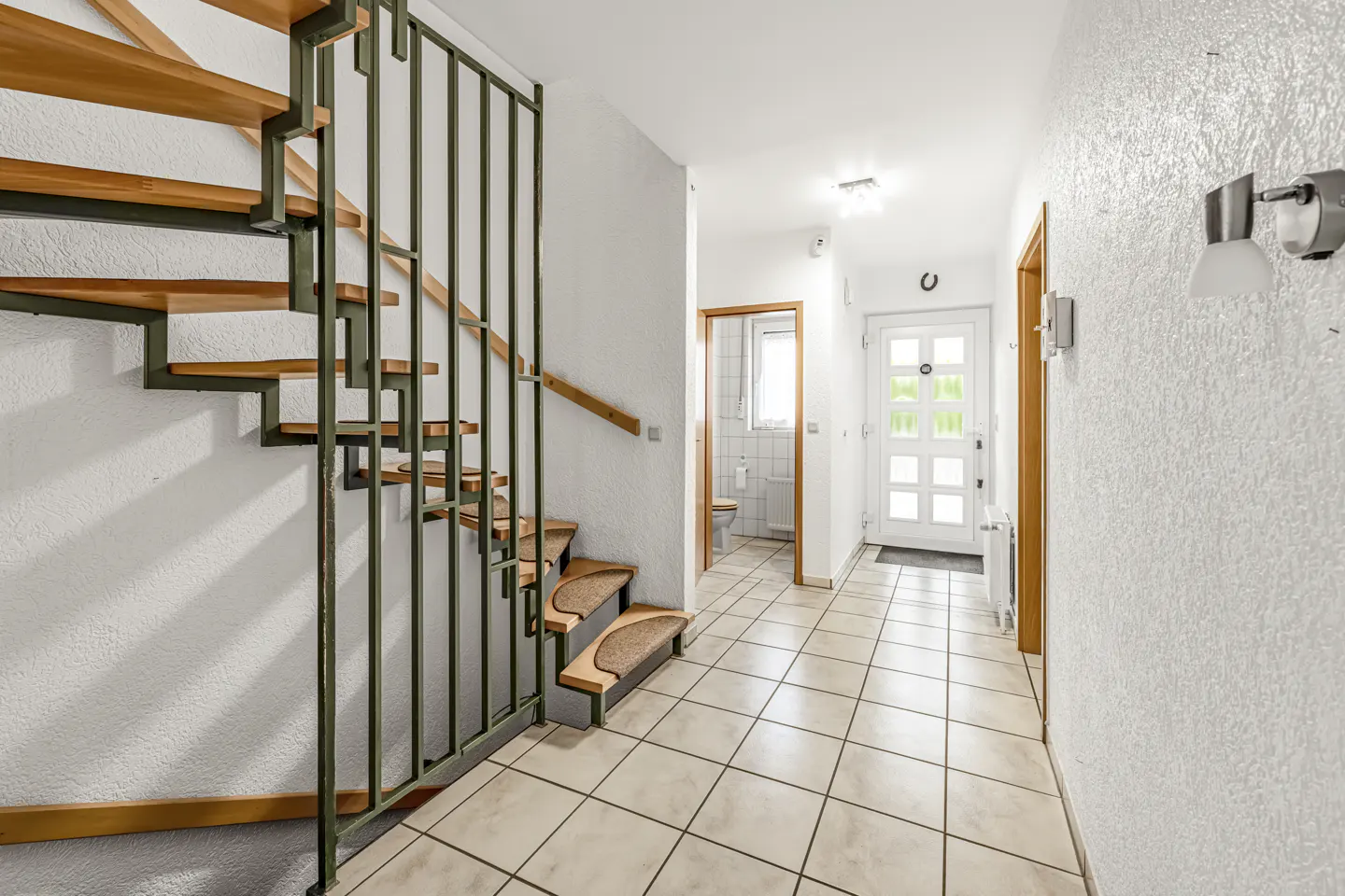 Bright hallway with tiled floor, wooden staircase with green metal railing, white front door, and a view into a bathroom.