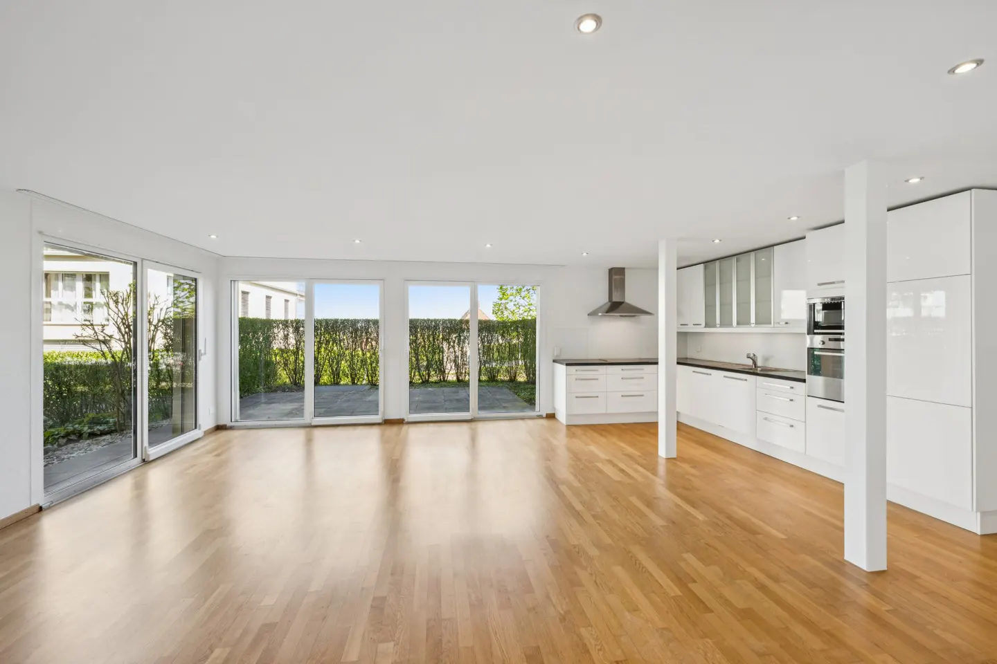 Bright, open-concept living space with hardwood floors, white walls, and a modern white kitchen. Sliding glass doors lead to a patio with green hedges.