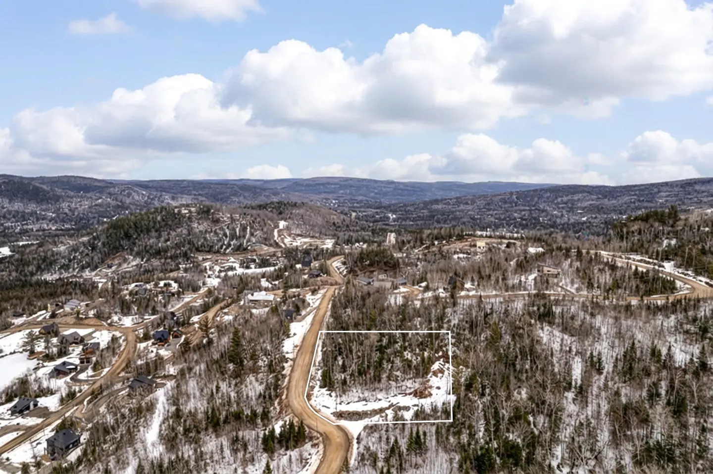 Aerial view of a wooded lot outlined in white, nestled in a snowy, hilly landscape under a cloudy blue sky.