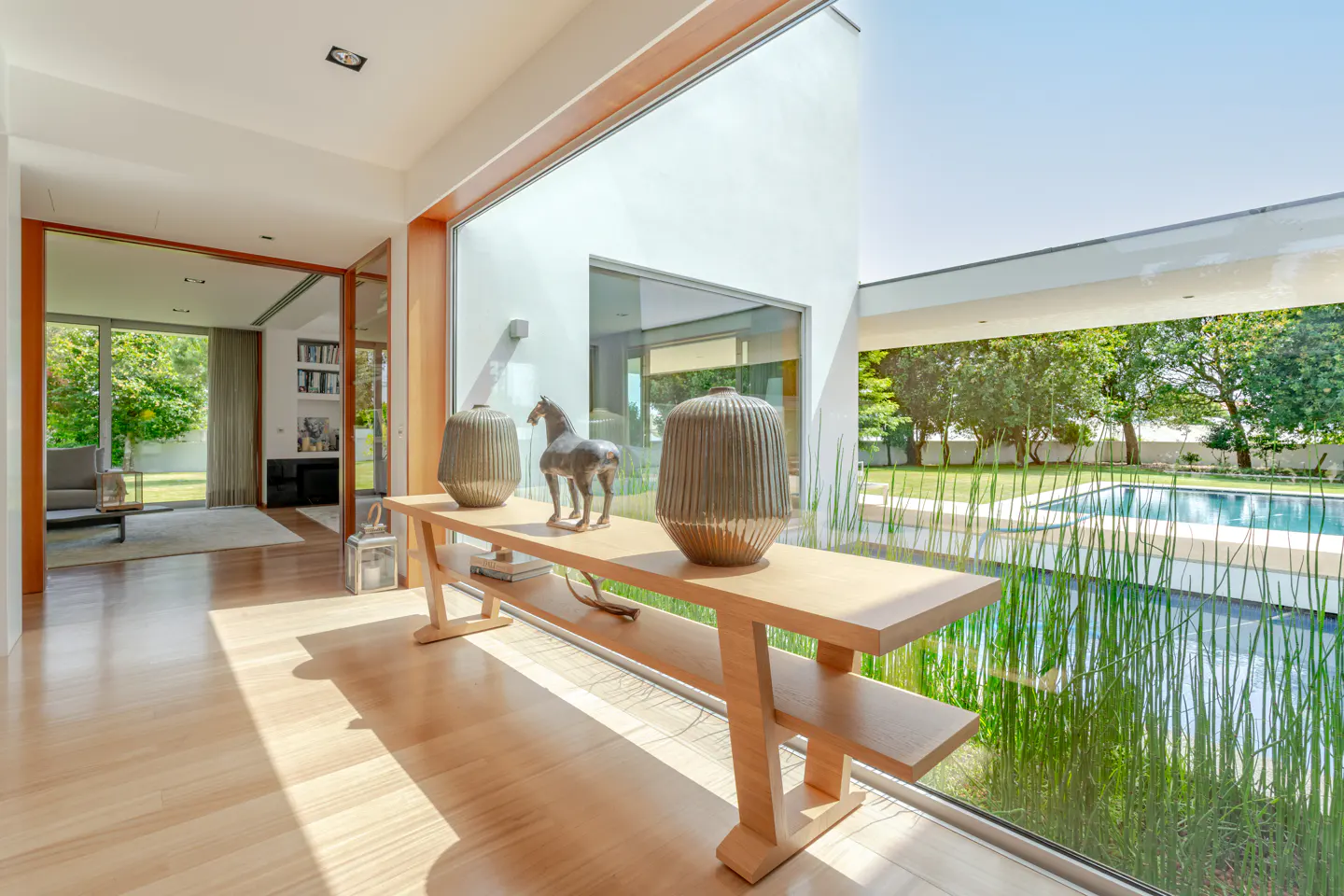 Bright foyer with wood floors, a console table with vases and a horse statue. A pool and greenery are visible through a large window.
