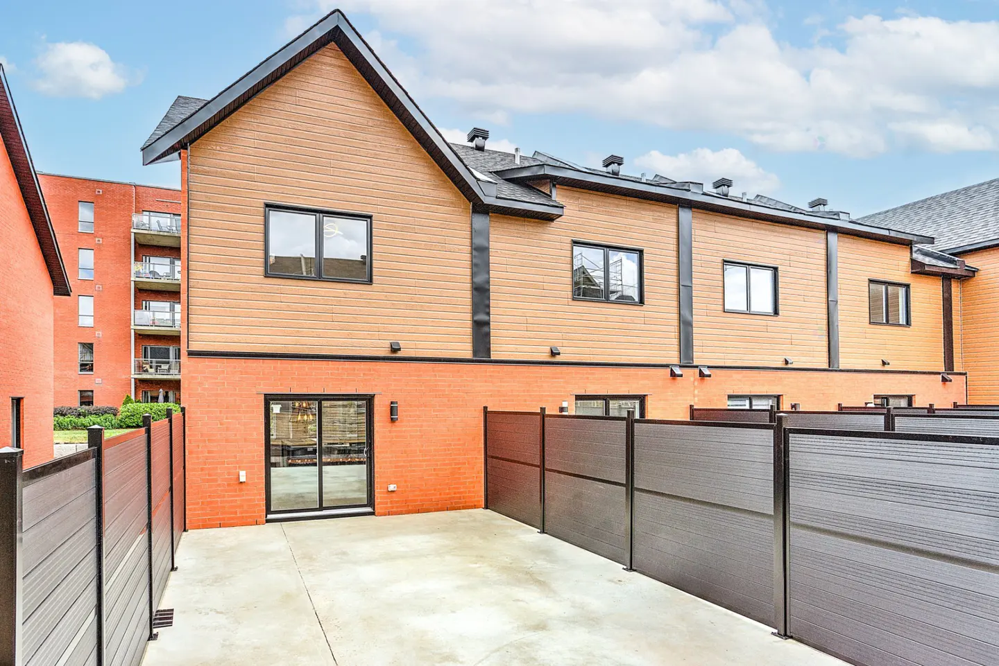 Exterior view of a two-story townhouse with a fenced-in patio. The building has a brick base and tan siding, with black window frames and trim.