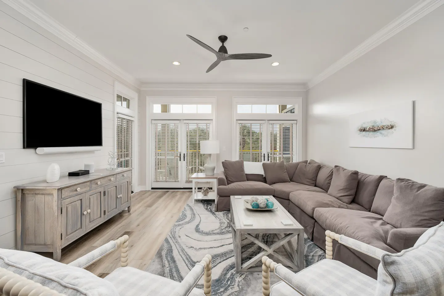 Bright living room with a gray sectional sofa, a TV, and a ceiling fan. Double doors lead to a balcony. A gray and white rug is on the floor.