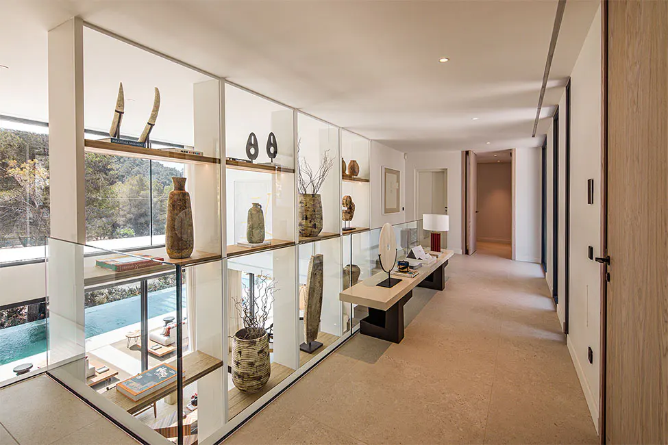 Hallway with glass railing overlooking a pool. Shelves display vases and art. A console table with a lamp sits along the wall.