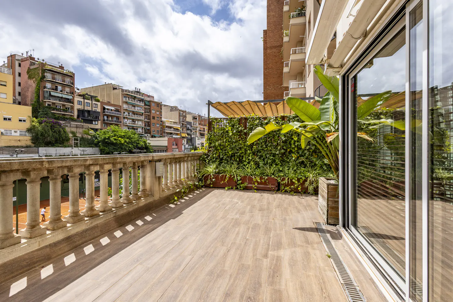 A wooden terrace with a stone balustrade overlooking a tennis court, featuring a green privacy wall and city buildings.