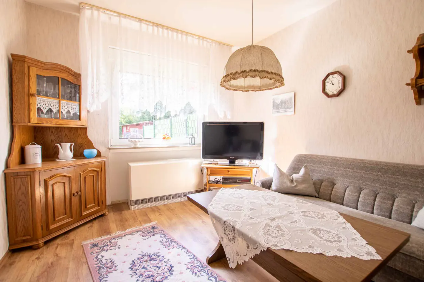 Cozy living room with wood floors, a gray sofa, and a wooden coffee table with a lace tablecloth. A TV sits near a window with sheer curtains.