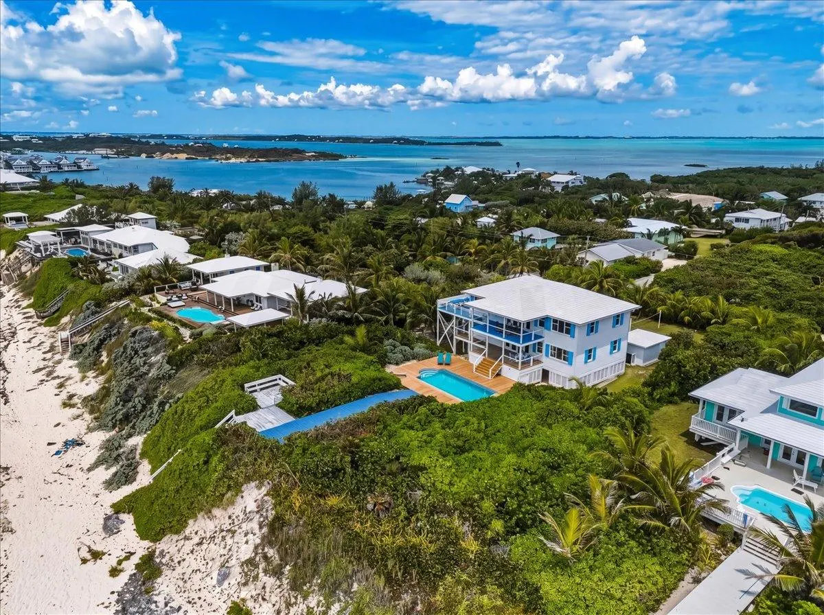 Aerial view of white houses with pools surrounded by lush greenery, with ocean and blue sky in the background.