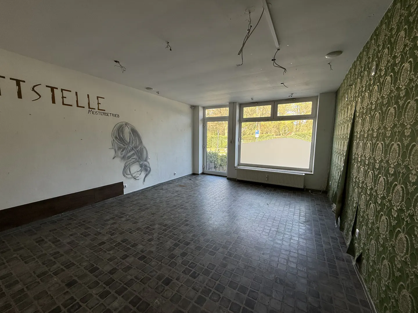 An empty room with gray tiled floors, white walls with graffiti, and green patterned wallpaper. A window shows trees outside.